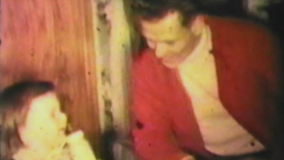 George Carroll wearing a red jacket sitting next to one of his children holding a bottle as they smile at each other.