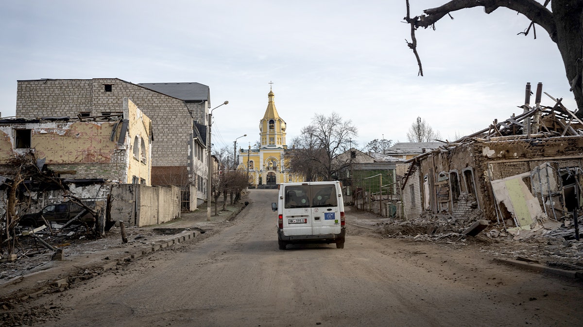 A lone evacuation vehicle travels along a damaged street in a heavily ruined Ukrainian city.