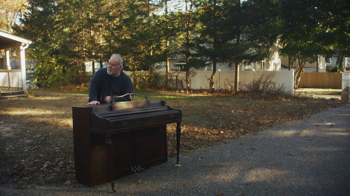 Mike Carroll stands outside his house in the daytime, leaning on the piano that's been moved to the end of the driveway.