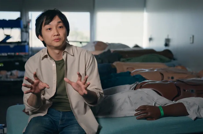 A man sits on the edge of a hospital bed with a row of partially clad manequins lying in a row behind him with their buttocks visible.