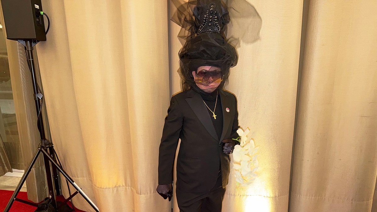 Andre Soriano Atelier dressed in an all-black formal suit with an oversized sculptural headpiece, holding white flowers at "Melania" premiere.