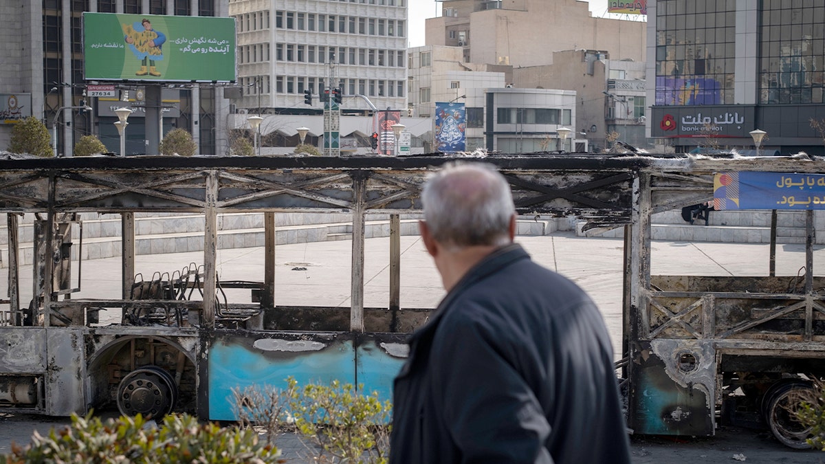 A man examines the charred remains of a public bus placed in an open plaza.