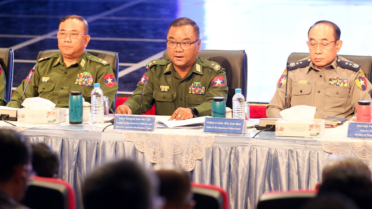 A government official speaks at a podium during a briefing, with microphones and a backdrop behind him.