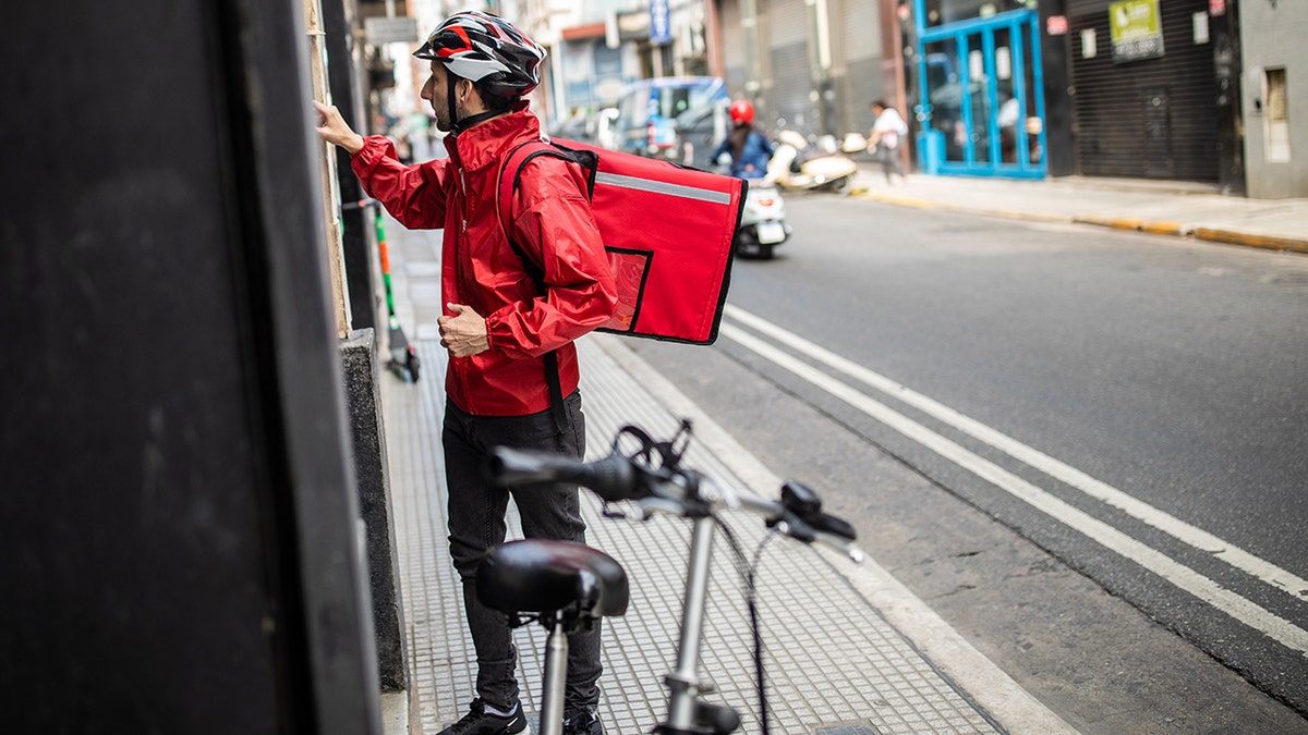 Pizza delivery guy knocking on bike with food backpack on back, helmet, and his bike next to him on city street.