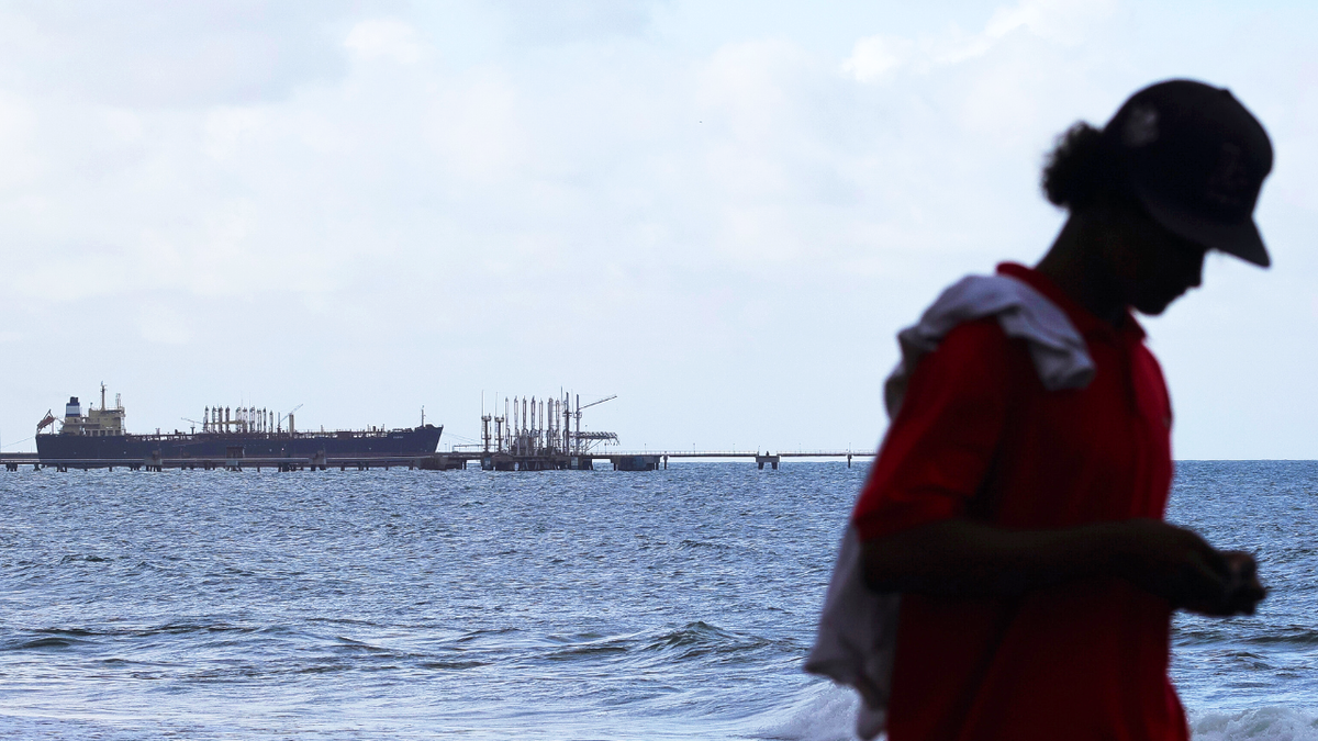 A man is seen walking along the beach while an oil tanker sits at a port.