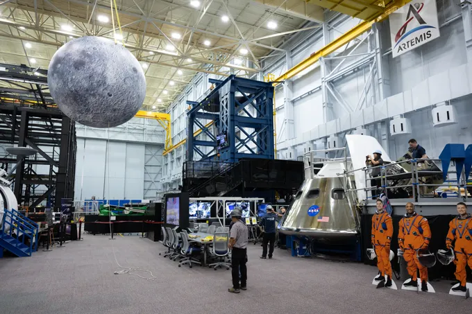 A large hangar-like building with a big inflatable moon hanging in the upper right corner and a model spacecraft, a row of computers and three cardboard cutouts of astronauts on the floor.