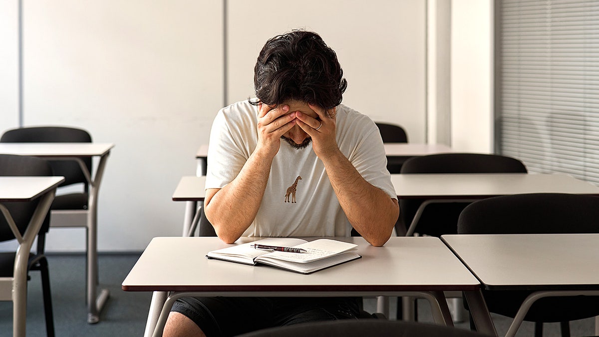 A young male student sits at a desk in a classroom with his face buried in his hands, appearing stressed and overwhelmed.