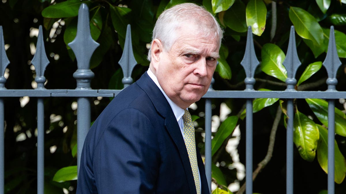 Ex-Prince Andrew in a dark blue suit and yellow tie walking in front of a fence.