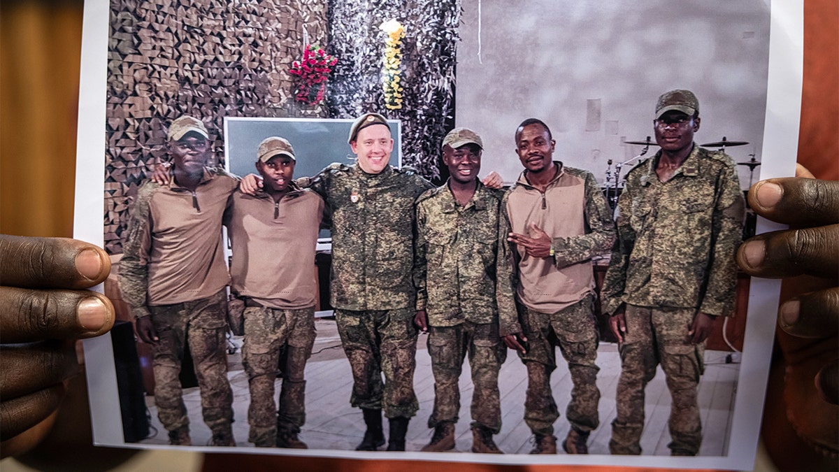 A man displays a printed photograph of several uniformed soldiers while standing indoors.