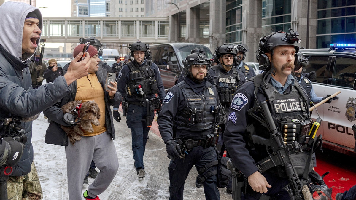 Police officers in protective gear move into a downtown street as a crowd gathers nearby.