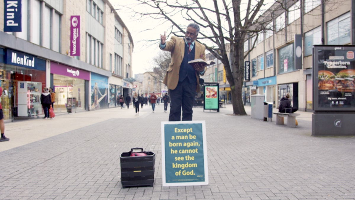 Dia Moodley standing on a platform and preaching on a city sidewalk next to a religious sign.