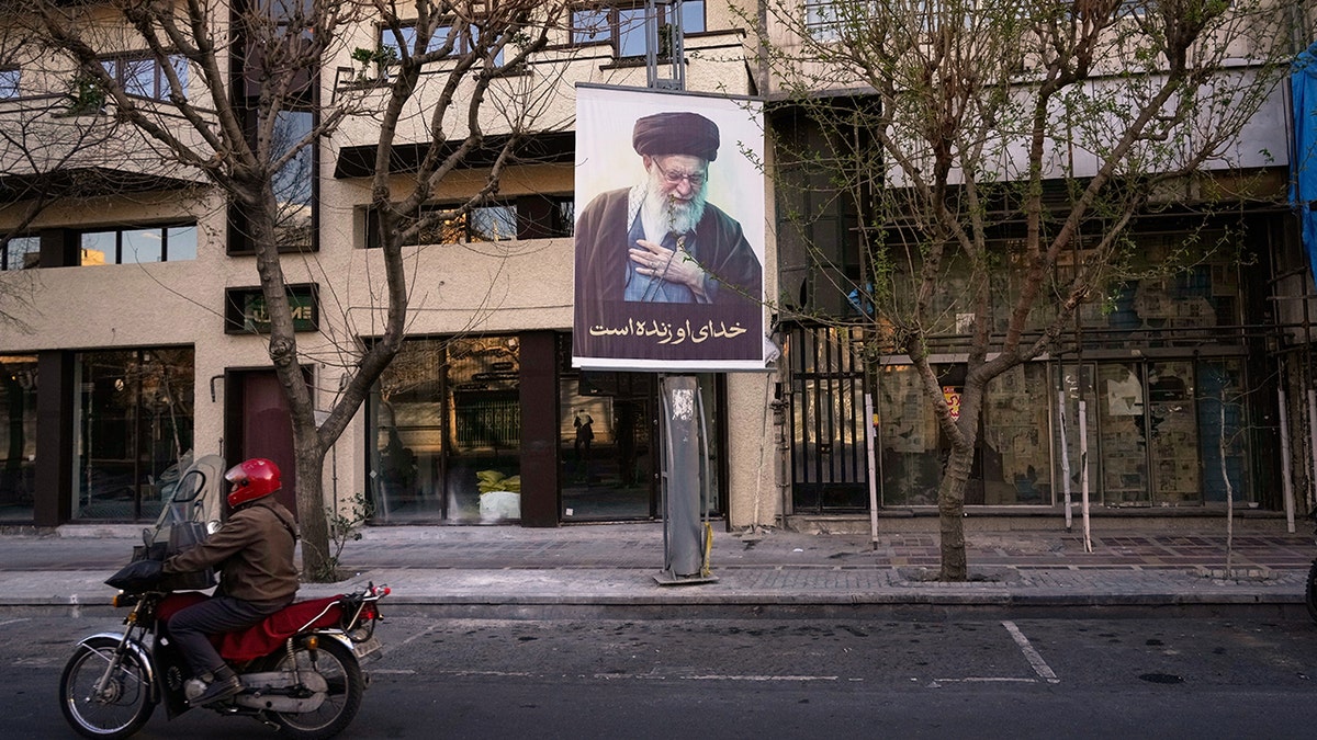 A lone motorcycle driving down a wide, deserted street in Tehran past a large portrait of Ayatollah Ali Khamenei.