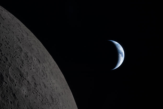 The cratered curve of the moon is in the foreground wiht a blueish crescent of Earth in the background.