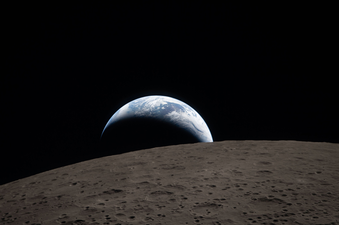 The curved surface of the moon in the foreground looks like a rich brown, not its more familiar gray. A blue and white crescent of earth rises in the distance.