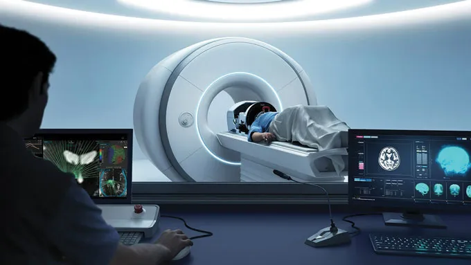 The view of an MRI scanner from the control room, with a person laying on the bed attached to the scanner, and a worker examining images on a screen.
