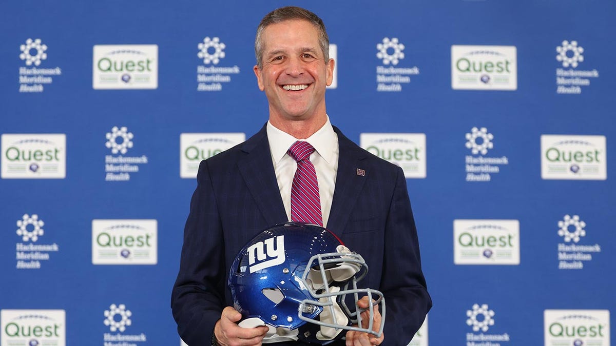John Harbaugh posing with a New York Giants helmet at a press conference