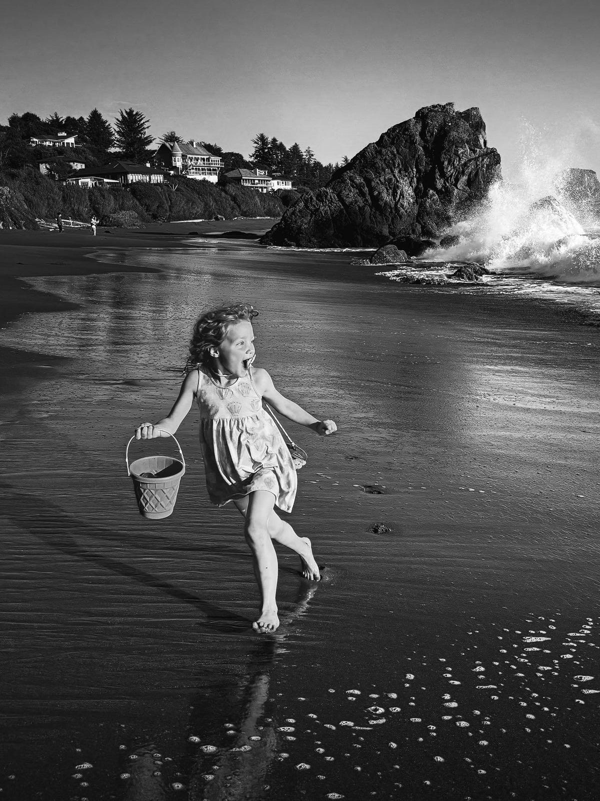 A young girl a beach carrying a bucket runs away from the incoming water.