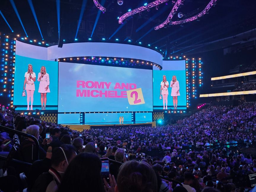 Two women appear on stage in front of a screen with the words "Romy And Michele 2" in front of a crowd of people.