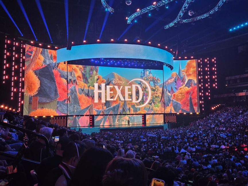A large screen with the words "Disney Hexed" on it over an animated colorful European village in front of a large crowd of people.