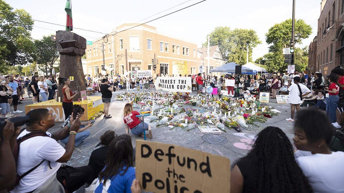 People gather around the Corey Ruiz protest encampment in Madison, Wisconsin, with a raised-fist sculpture and protest signs visible.