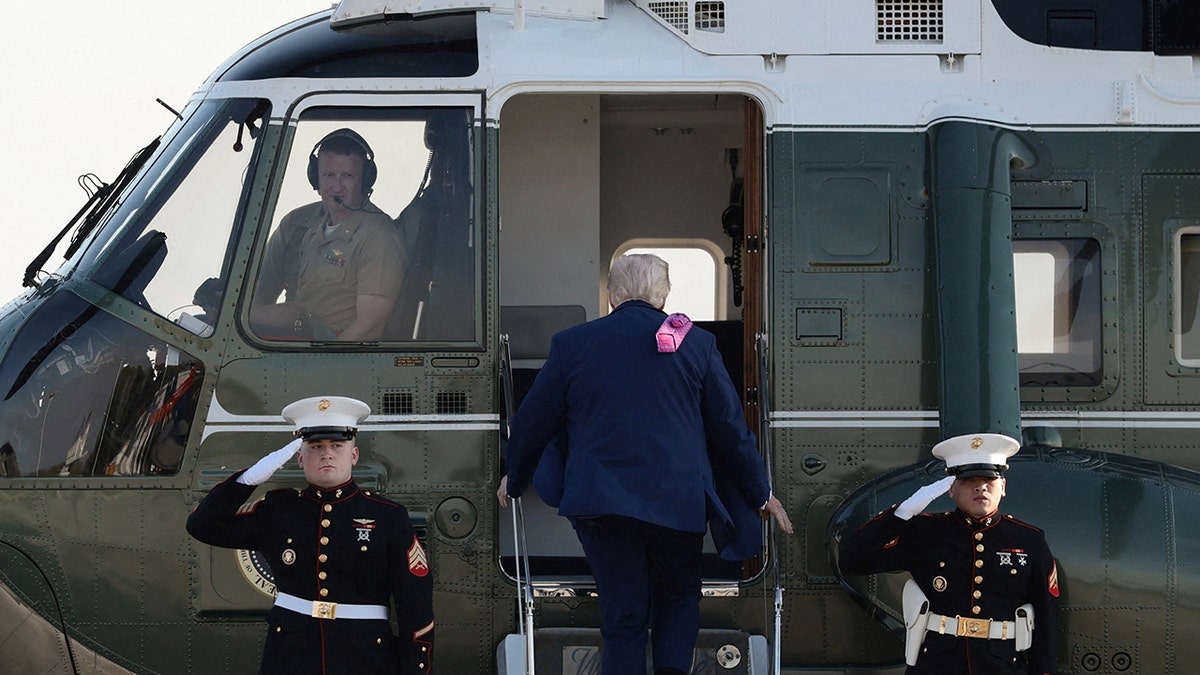 Donald Trump boarding a Marine One helicopter on a landing field.