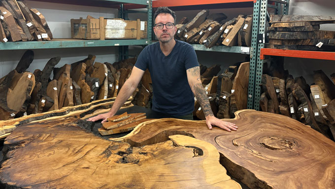 Kevin Anchukaitis stands behind large cross sections of trees at the University of Arizona’s Laboratory of Tree-Ring Research in Tucson, with shelves of wood samples behind him.