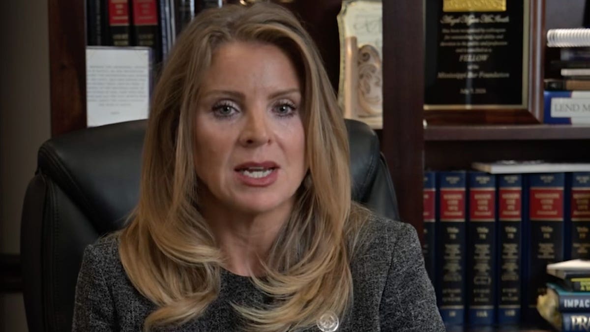 A woman with long blond hair in a gray tweed blazer speaks while seated at a desk in an office lined with legal books and framed awards.