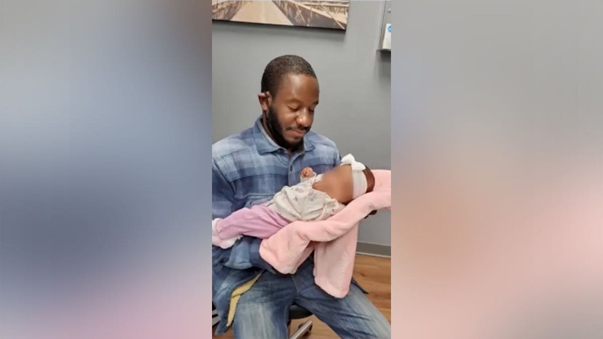 A man in a plaid shirt sits and cradles a sleeping infant wrapped in a pink blanket.