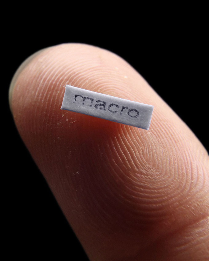 A tiny piece of paper with the word "macro" on it rests on a pointer finger. The paper is smaller than the pad of the finger.