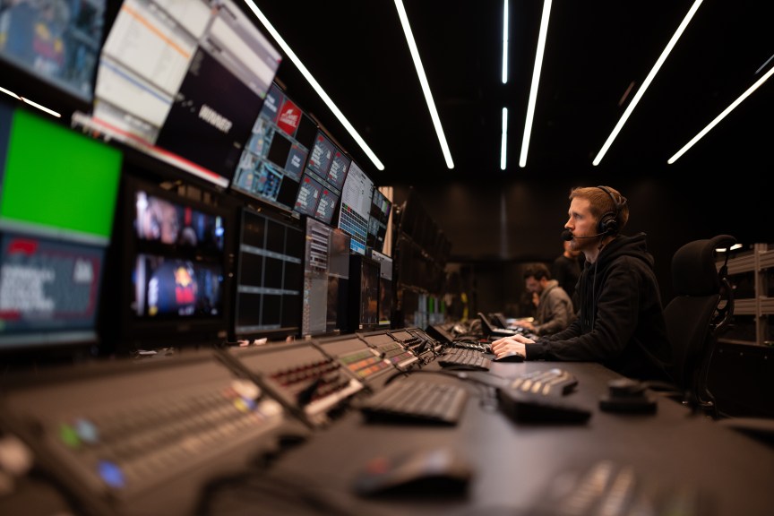 A person looks up at the screen at the end of a row of monitors and switchboards
