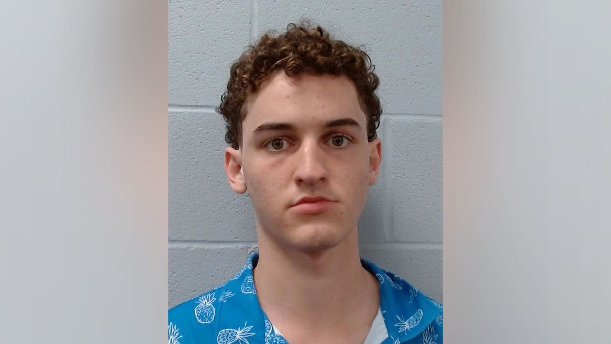 Booking photo of Tyler Fogg wearing a blue pineapple-patterned shirt against a gray cinder block wall.