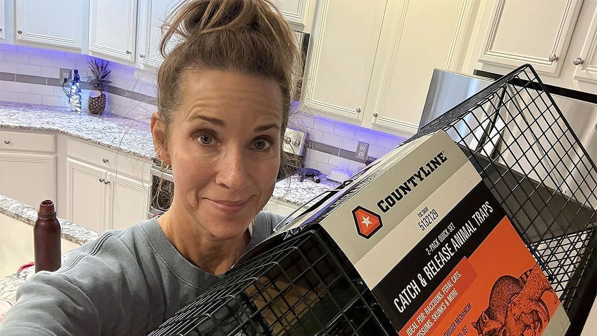 Lynda Loveland poses in a kitchen while holding a large black wire animal trap.