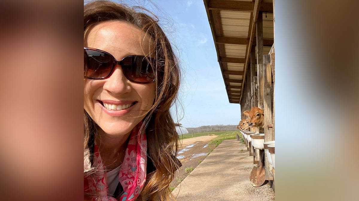 Lynda Loveland smiles while wearing sunglasses near a row of cattle feeding inside a barn.