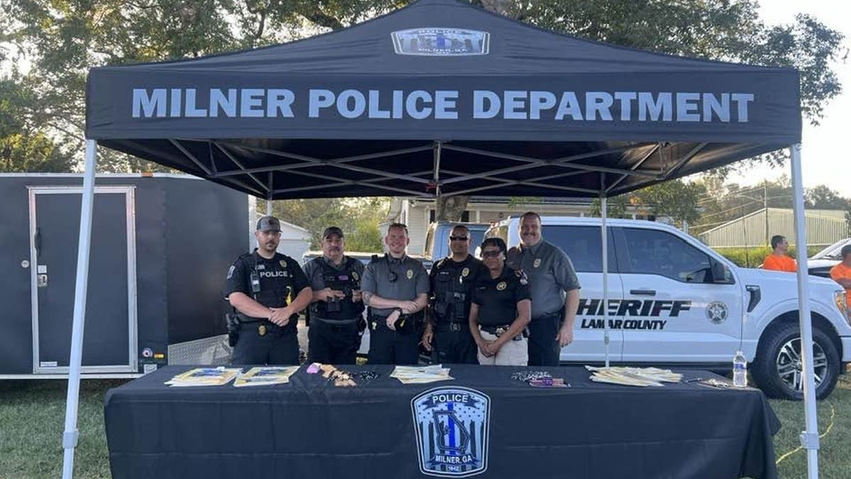 Six people pose beneath a Milner Police Department tent at an outdoor event, with a Lamar County Sheriff's Office vehicle parked behind them.
