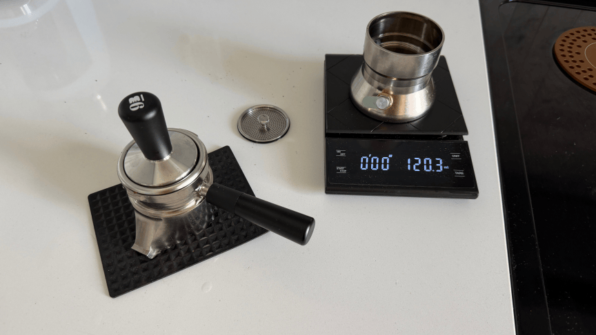 The bottom section of a 9Barista espresso maker sits on a kitchen scale, while espresso grounds are tamped into the upper part on a white countertop.