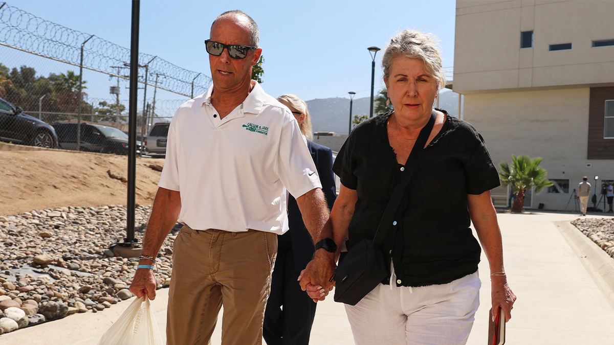 Martin Robinson, wearing sunglasses and a white polo shirt, holds hands with Debra Robinson outside a building